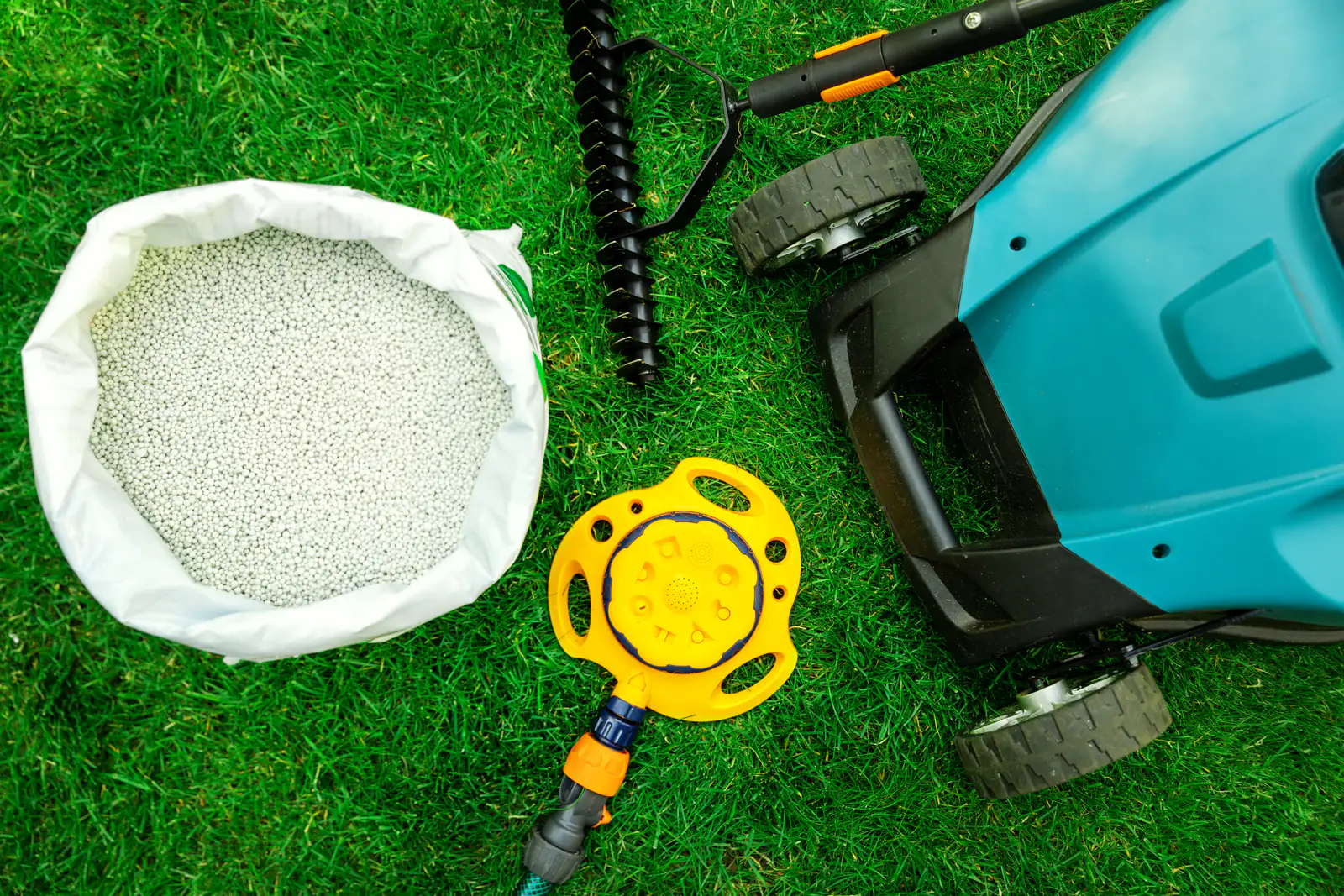 Summer lawn care essentials on a freshly mown green lawn: an open bag of slow-release fertilizer, a core aerator, a lawn sprinkler and a push mower